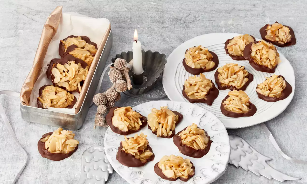 Florentiner mit goldbraunen Mandelblättchen und glatter dunkler Schokoladenschicht arrangiert auf Tellern und in einer Metallbox mit Papier