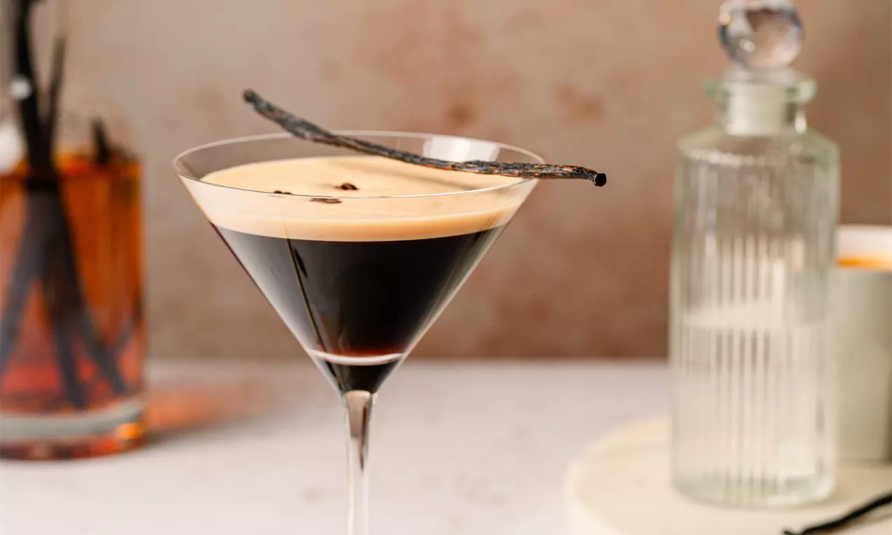Picture - Dark espresso martini in a clear, sleek glass with creamy foam top, garnished with coffee beans and a vanilla pod on a smooth white coaster
