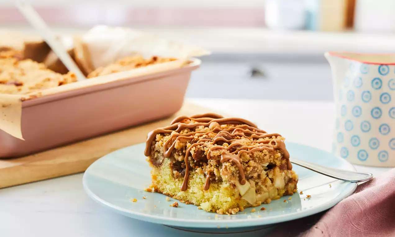 A slice of apple and Biscoff crumble cake with golden layers, crumbly topping, and caramel drizzle on a pale blue plate with a fork