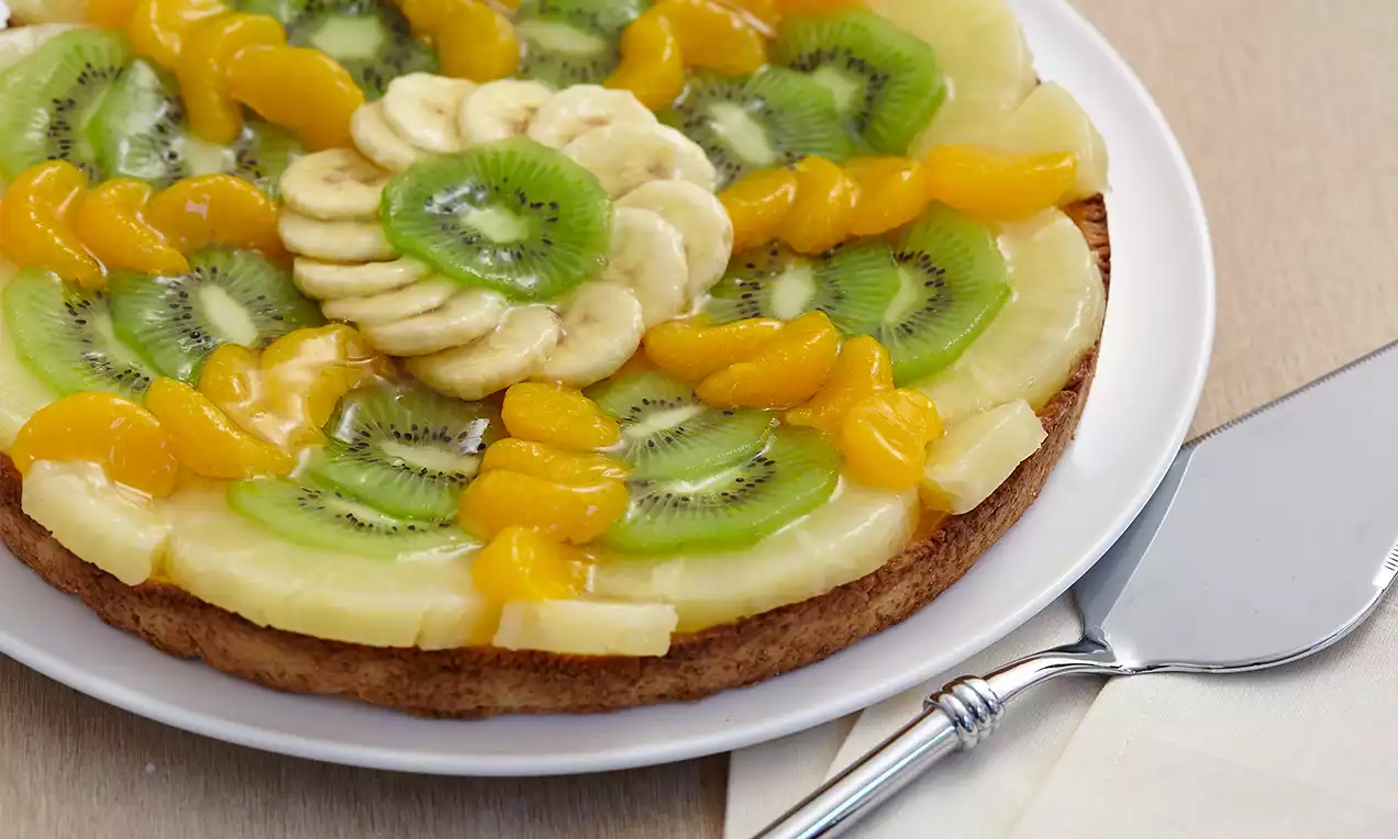 Früchtekuchen mit goldbraunem Boden, belegt mit gelben Mandarinenspalten, grünen Kiwi-Scheiben, hellen Bananen und Ananasringen auf weißem Teller