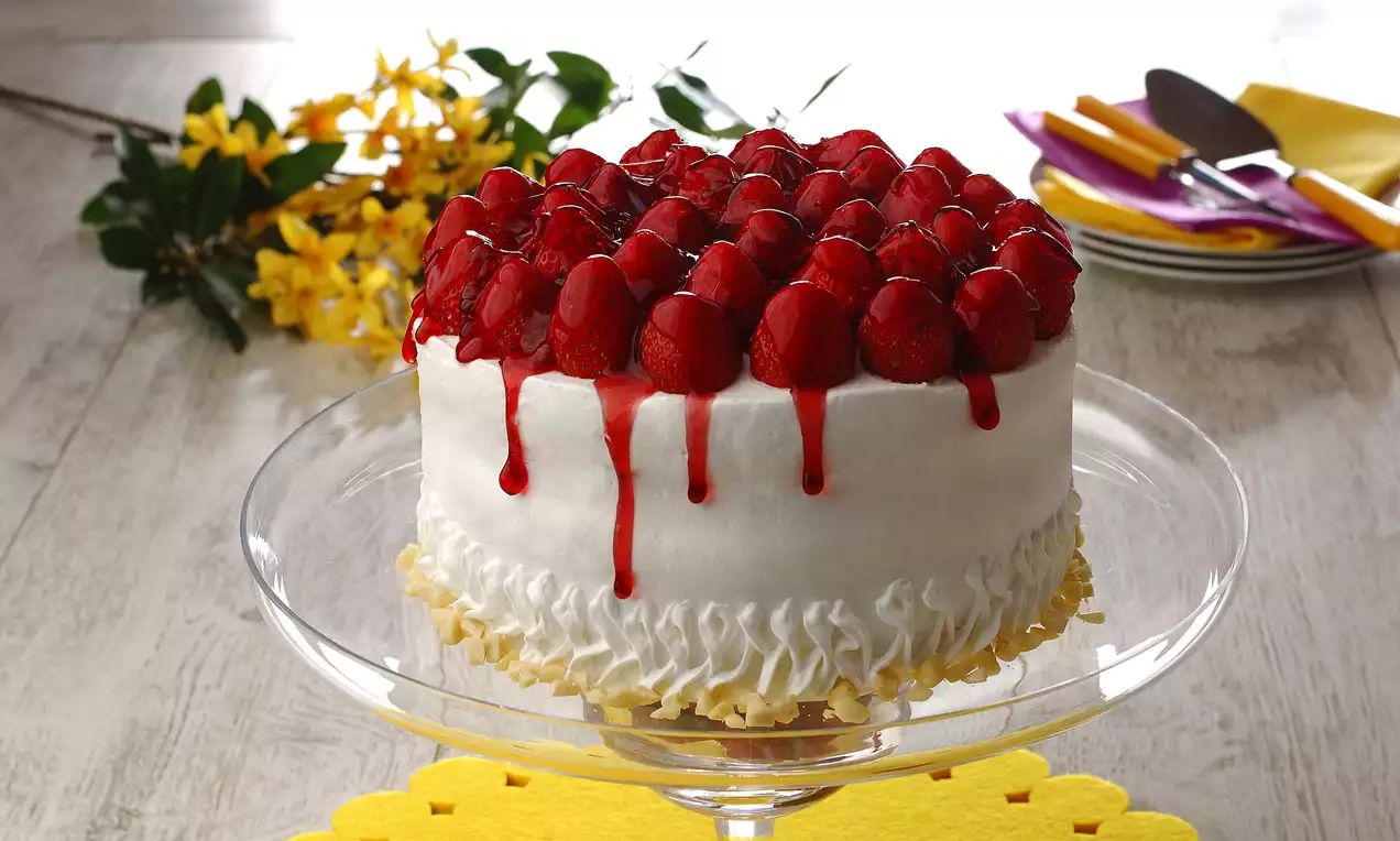 Üzerinde parlak kırmızı çilekler ve akışkan çilek sosu bulunan beyaz kremalı, alt kısmı kabartmalı, şeffaf ayaklı tabakta sunulan pasta