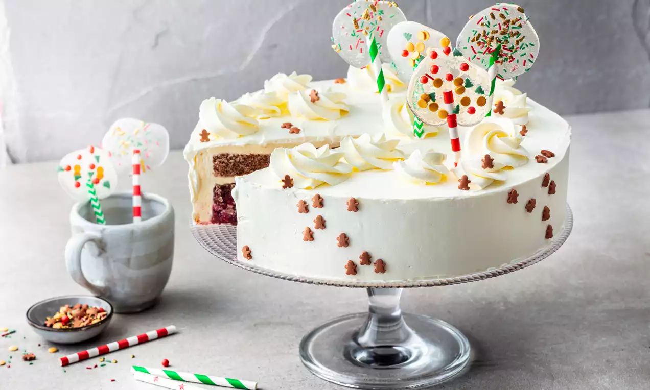 Witte botercrème taart met kleurrijke lollydecoraties, slagroomrozetten en kerstvormige strooisels op een glazen taartplateau