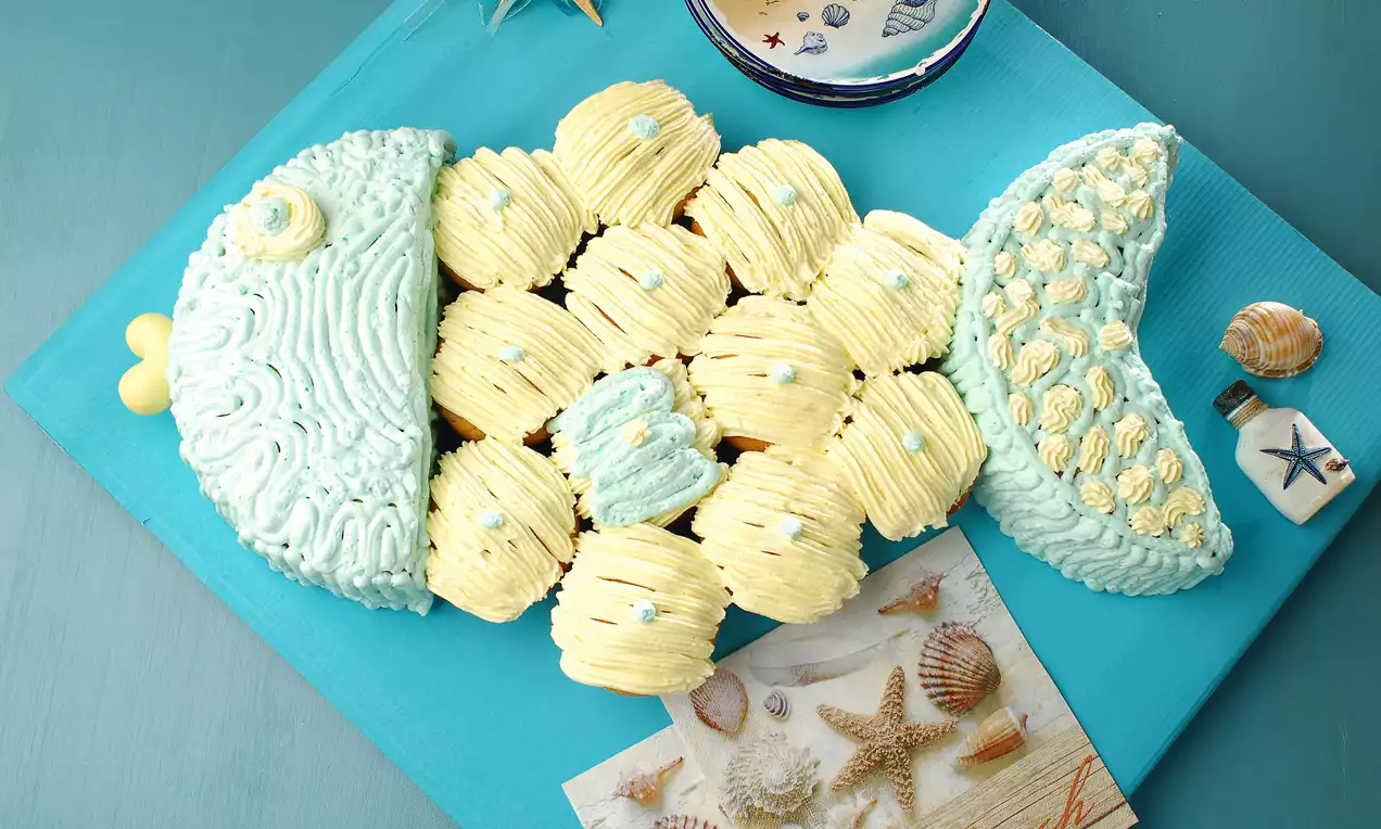 Vanilya ve açık mavi krema detaylı, balık şekilli pastel tonlarda pasta, deniz temalı dekorlarla mavi bir zeminde sunulmuş