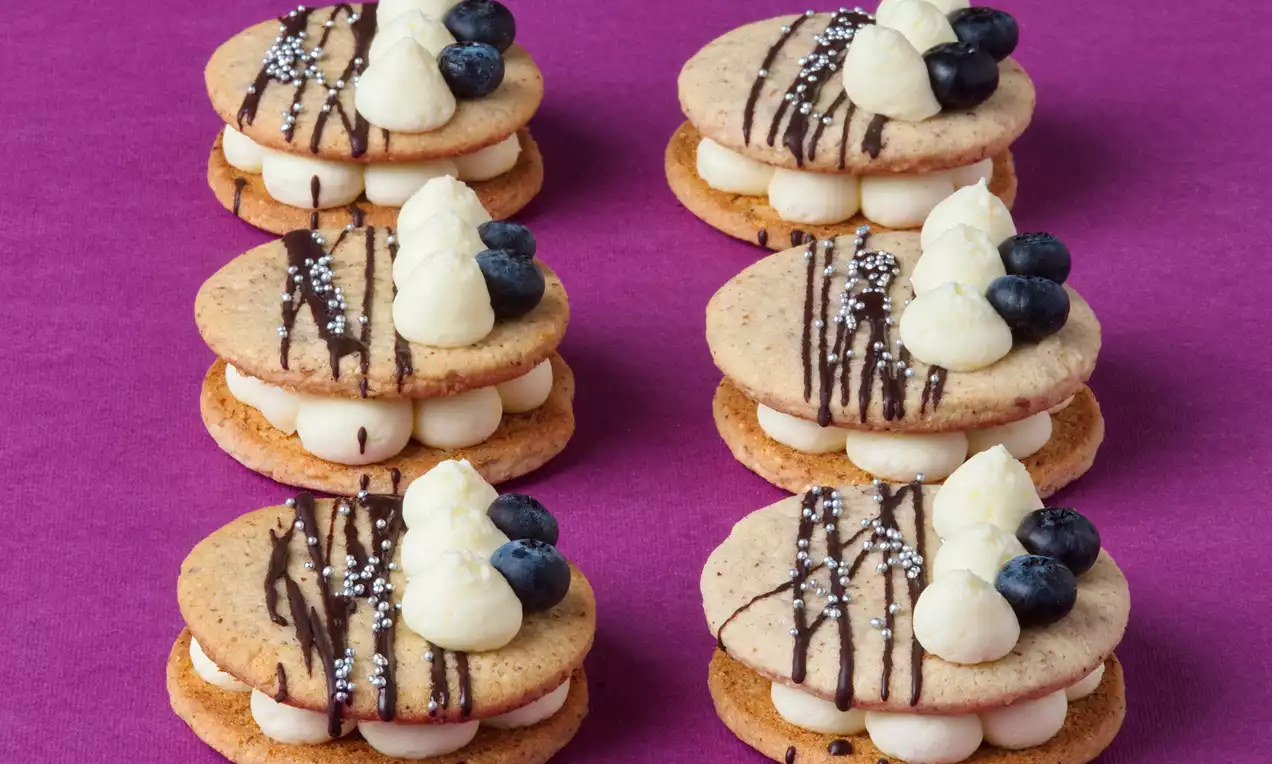 Kulaté krémové dortíčky zlatohnědé barvy, zdobené bílým krémem, čerstvými borůvkami a čokoládovými proužky, na fialovém pozadí