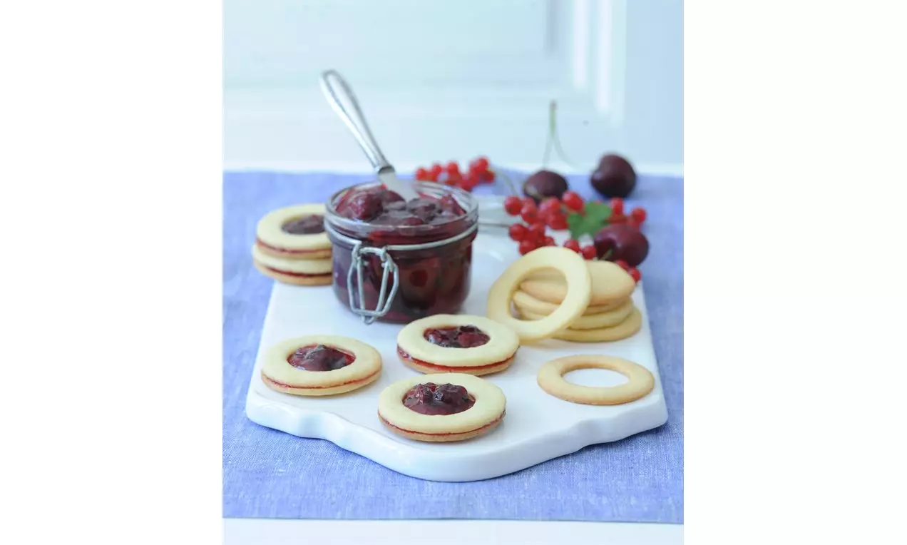 Světle zlaté linecké sušenky srdcovitého tvaru a lesklým červeným rybízovým džemem uprostřed na bílém podnosu vedle sklenice džemu a čerstvého ovoce