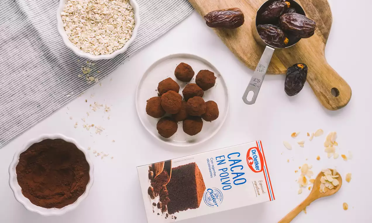 Trufas redondas cubiertas de cacao oscuro sobre un plato blanco acompañadas de dátiles, avena y nueces dispuestas en una mesa clara