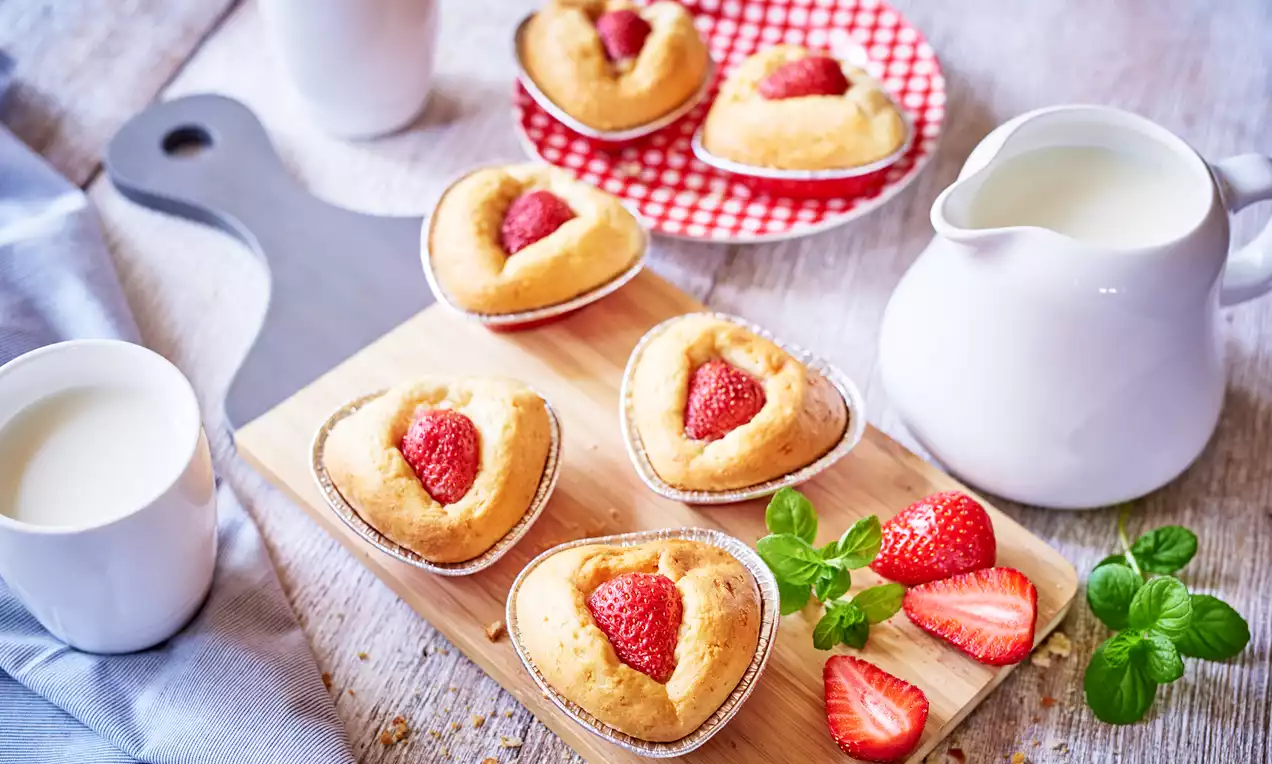 Babeczki w kształcie serc, z miękkiego, złocistego ciasta ozdobionej soczystymi, czerwonymi połówkami truskawek na drewnianej desce