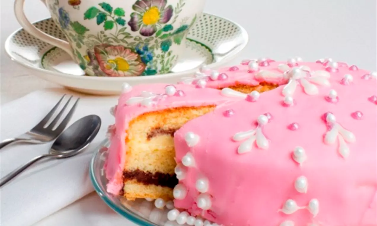 Rosa festkake med vaniljekrem og sjokoladefyll, pyntet med hvite perler og glasurblomster, servert på glassfat med kopp i blomstrete mønster