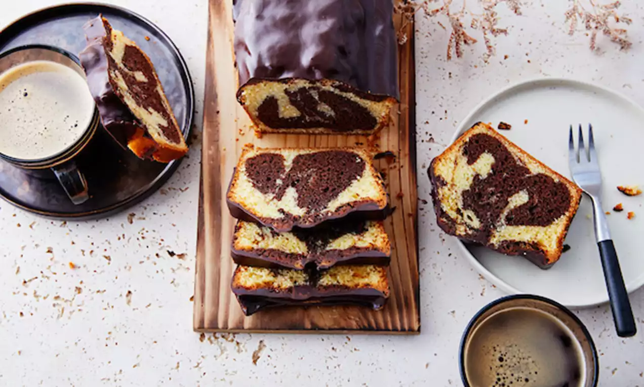 Marmercake met goudbruine korst en donkere chocoladeglazuur, in plakken gesneden op een houten plank naast kopjes koffie en een wit bord