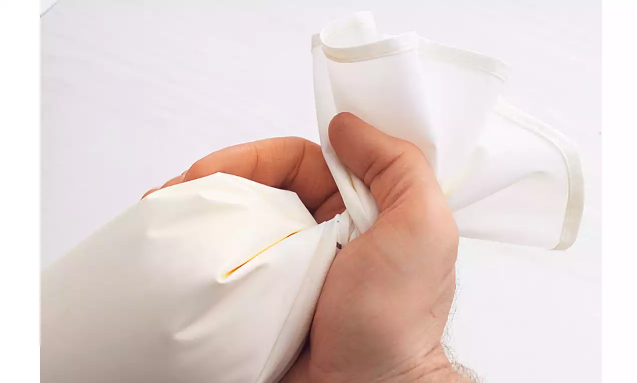 Eine Hand hält einen weißen Spritzbeutel am oberen Ende gefasst, während sich die gefüllte, glatte Oberfläche leicht wölbt