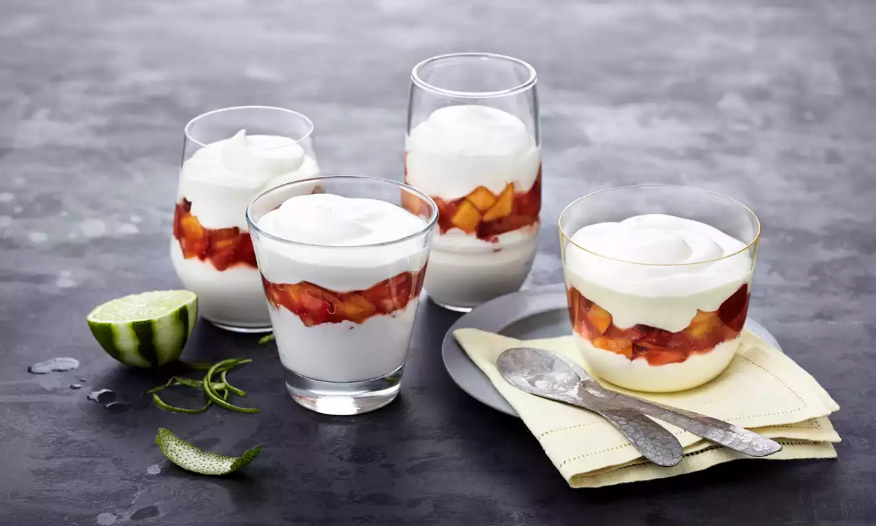 Gläser mit weißer Quarkspeise, Schicht aus rot-gelbem Obst, daneben Limettenschnitze und -zesten sowie Besteck auf gelber Serviette