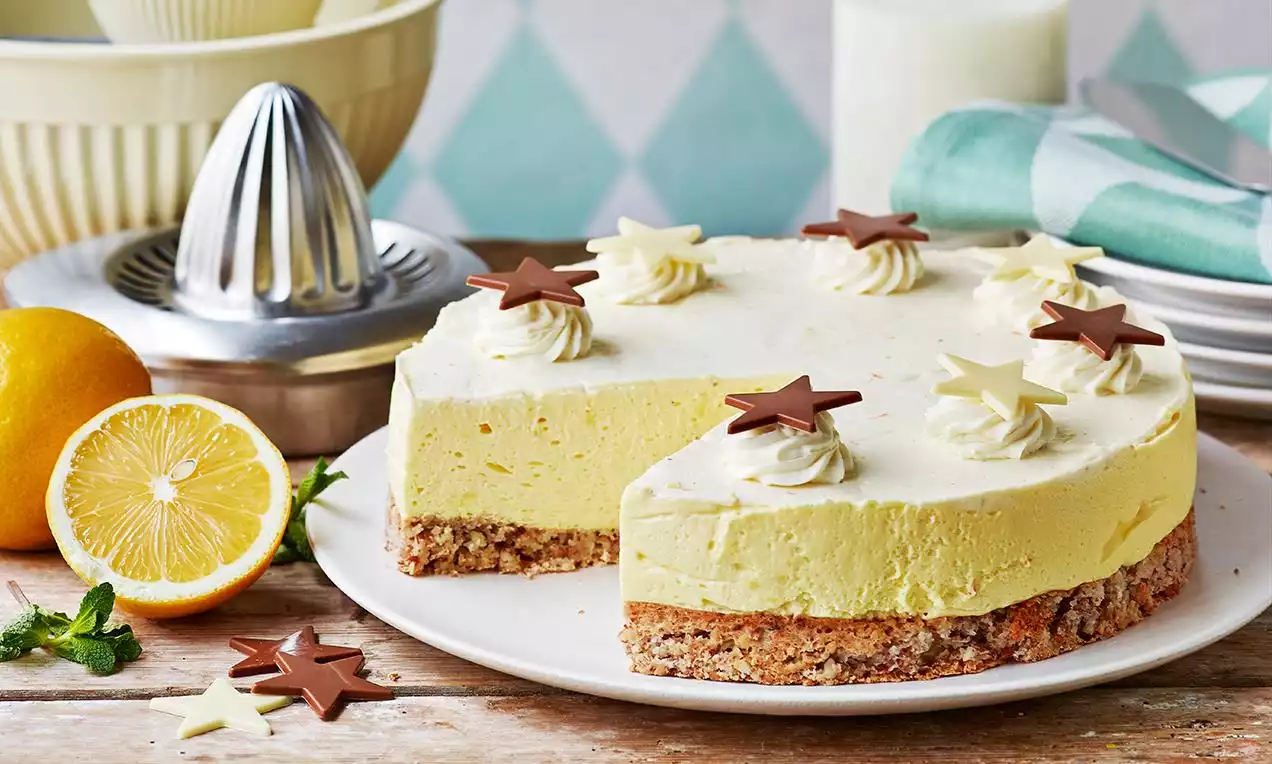Sitronmousse med lys gul krem på sprø mandelbunn, pyntet med hvite kremtopper og sjokoladestjerner på hvitt fat ved siden av sitron og mynteblad