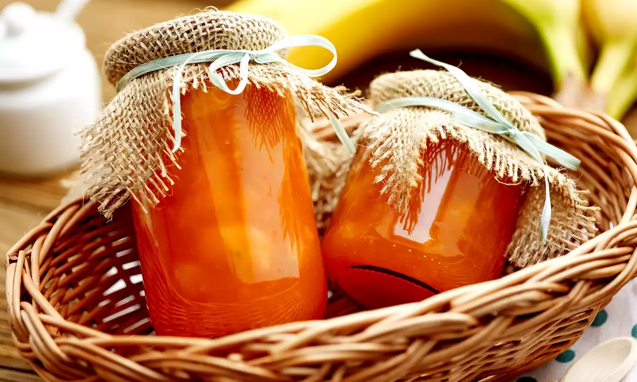 Glasheldere potten met oranje bananenconfituur, bedekt met strodeksel en lint, geplaatst in een gevlochten mand naast rijpe gele bananen