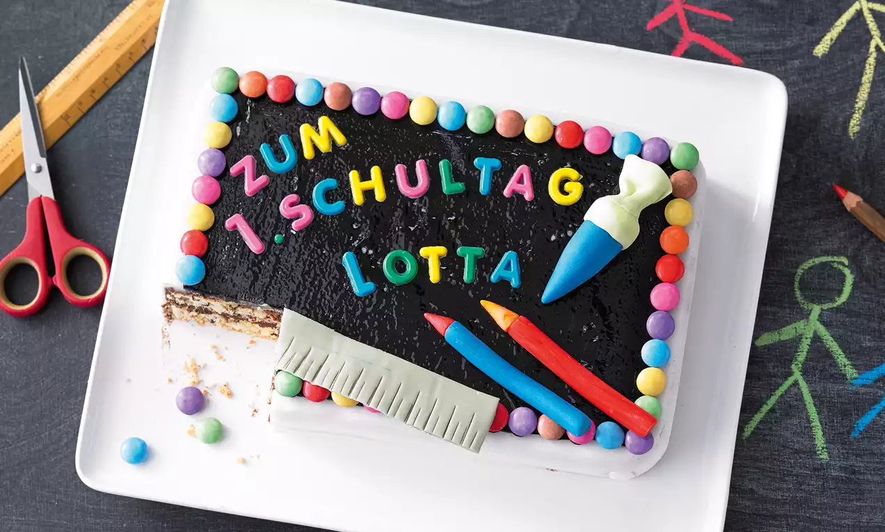 Schokoladen-Schultafel mit buntem Schriftzug, verziert mit essbaren Farbkreiden, Lineal und Zuckerkugeln auf einem weißen Teller