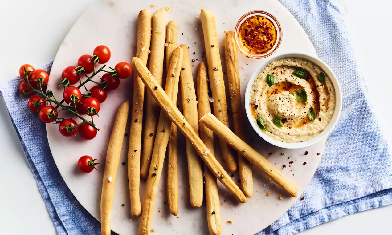 Knusprige, goldgelbe Breadsticks auf weißem Teller, dekoriert mit Kirschtomaten, cremigem Dip und Gewürzen auf einem blauen Tuch