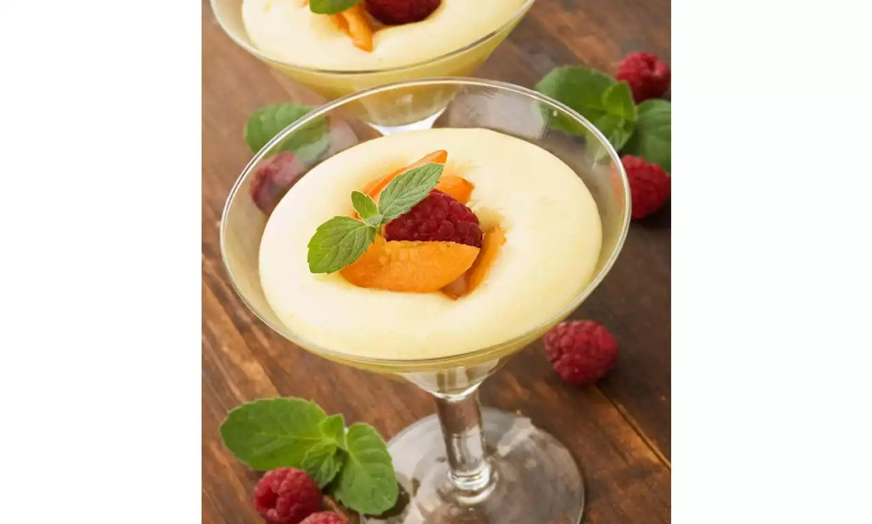 Lemon mousse in a clear glass, pale yellow and creamy, topped with a raspberry, orange slices, and fresh mint on a wooden table