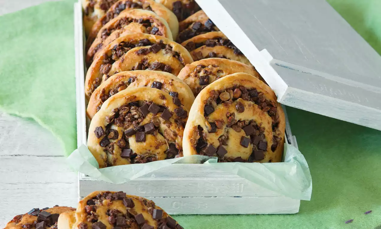 Zlatavé spirálky z listového těsta s vrstvenou náplní z křupavého müsli a tmavé čokolády, vyskládané v bílé dřevěné krabičce na zeleném podkladu