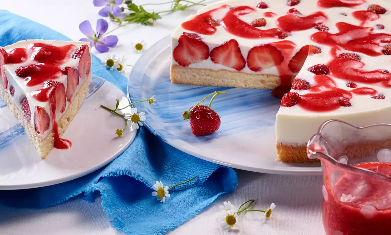 Joghurt-Panna-Cotta-Erdbeer-Torte mit weißer Sahnecreme, frischen Erdbeerscheiben und rotem Fruchtspiegel auf blauem Teller mit Blütendekoration