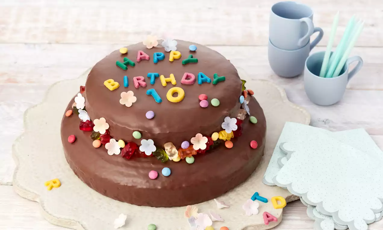 Runde Geburtstagstorte mit glatter Schokoglasur, bunt verzierten Lettern, kleinen Blüten und Zuckerdrops auf einem weißen Untersetzer
