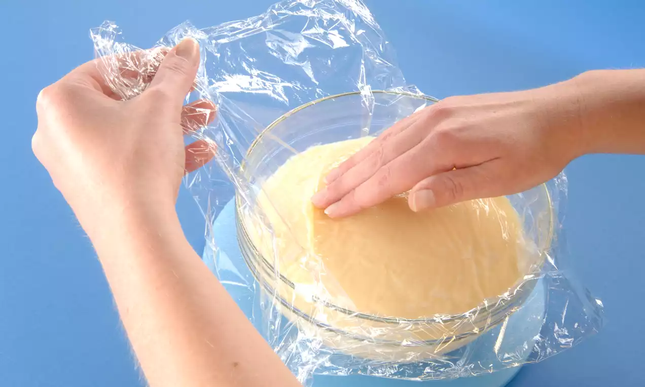 Hellgelber Pudding in einer durchsichtigen Schüssel, mit einer straff darüber gezogenen Frischhaltefolie bedeckt, Hände beim Andrücken der Folie