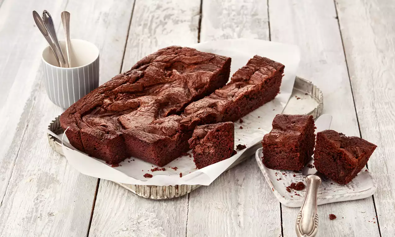 Tamnosmeđi brownie s bogatom, pomalo ispucalom korom, izrezan na nepravilne kocke, poslužen na bijelom papiru na srebrnom pladnju