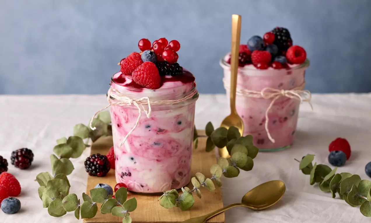 Gläser mit rosa Buttermilchcreme und Beerenmuster, dekoriert mit frischen roten und blauen Beeren, goldene Löffel und Eukalybusblätter