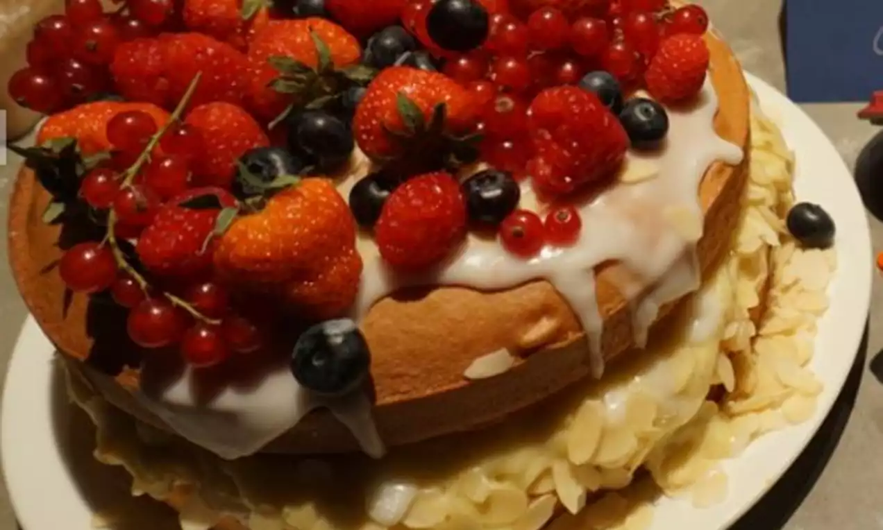 Warme, goudbruine laagjescake met ivoorkleurig glazuur, versierd met glanzend rode bessen, blauwe bosbessen en amandelschilfers op een wit bord