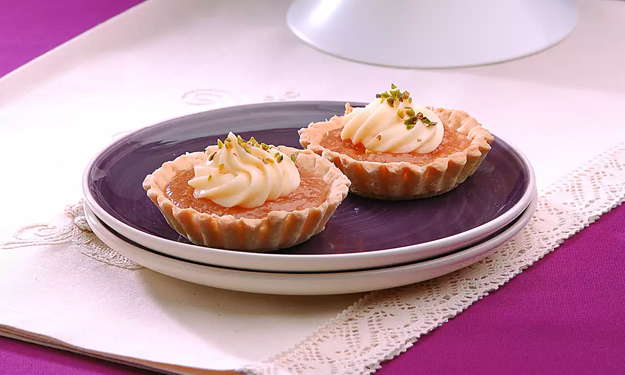 Altın rengi kıtır tabanlı armutlu tartoletler, kremsi beyaz dolgu ve ince kıyılmış yeşil Antep fıstığı ile mor tabakta sunulmuş