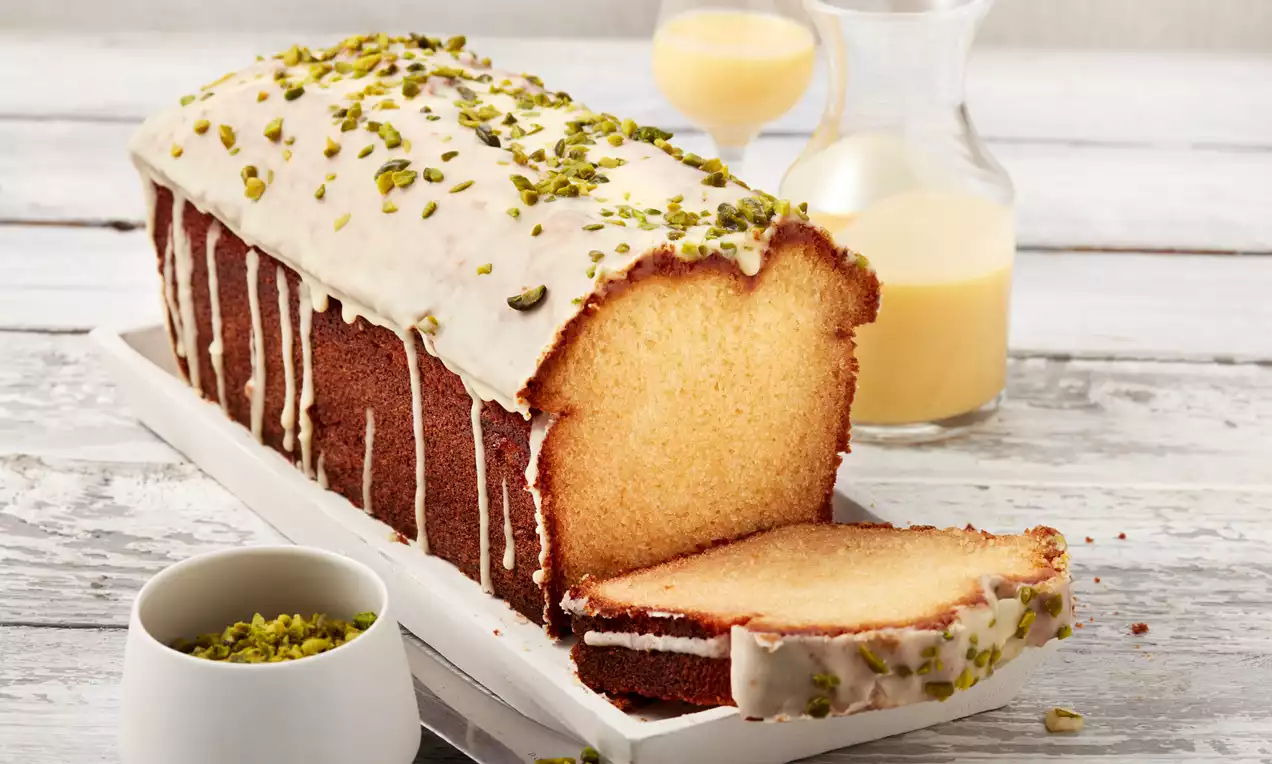 Saftiger, goldgelber Rührkuchen in Kastenkuchenform mit weißem Zuckerguss und gehackten Pistazien, angerichtet auf weißer Platte