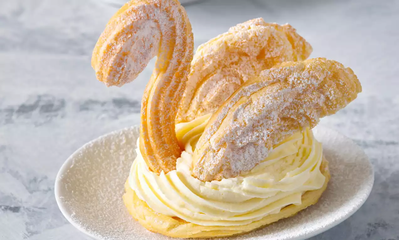 Labutě z odpalovaného těsta ve tvaru oblých křídel a krků, posypané moučkovým cukrem, na krémové náplni na světlém talíři