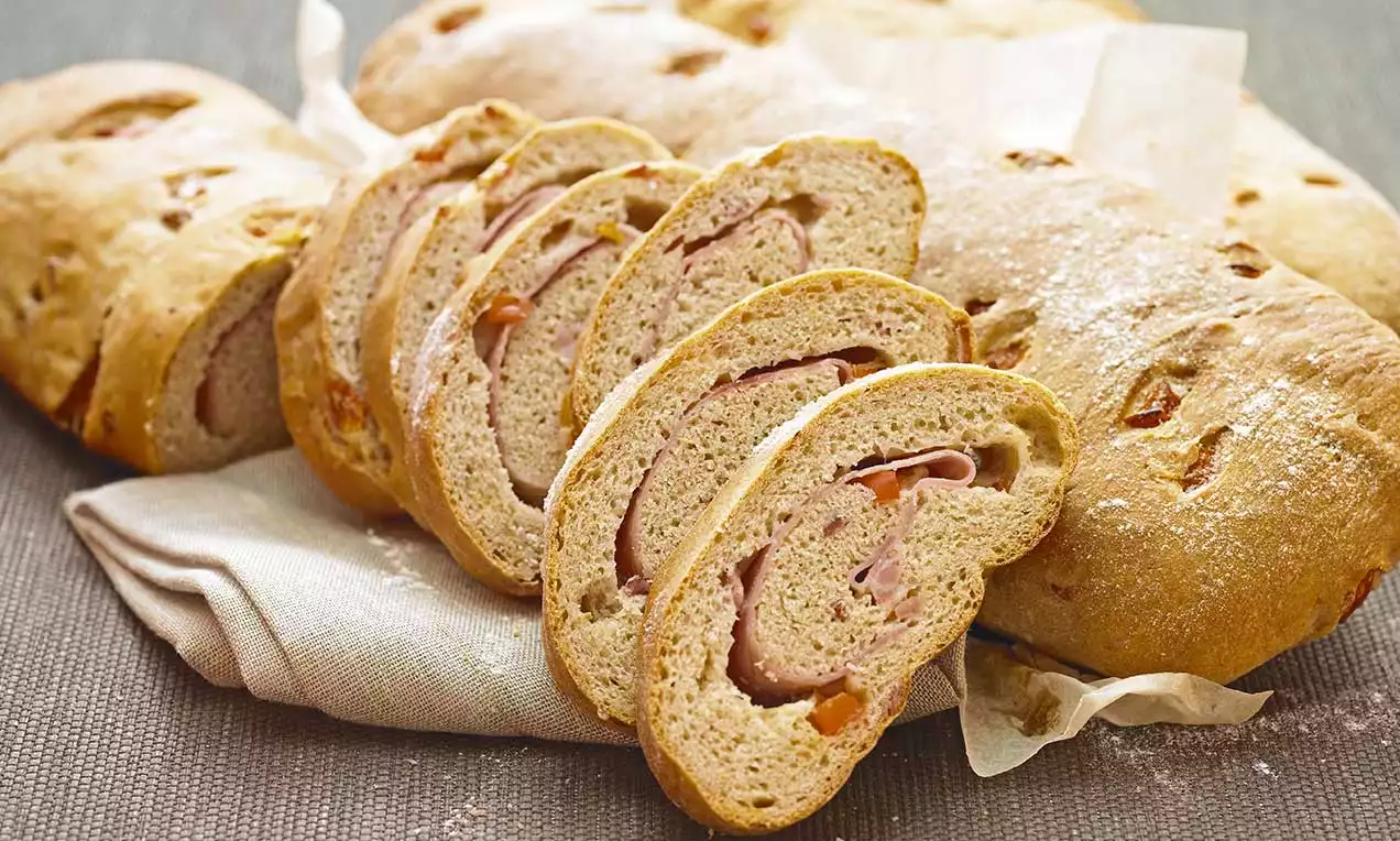 Scheiben von goldbraunem Brot mit lockerem Teig und sichtbarer Schinkenfüllung, arrangiert auf einem weißen Tuch mit ganzen Laiben im Hintergrund