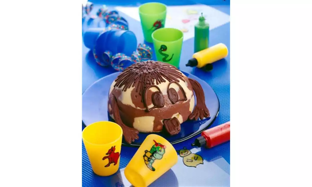 Pudding bicolore en forme de tête ronde avec des détails en chocolat dessiné sur une assiette bleue entourée de verres colorés