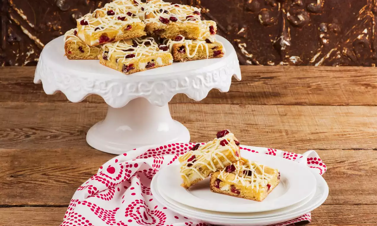Festive blondies golden and studded with crimson cranberries, drizzled with white chocolate, stacked on a scalloped white cake stand and plates