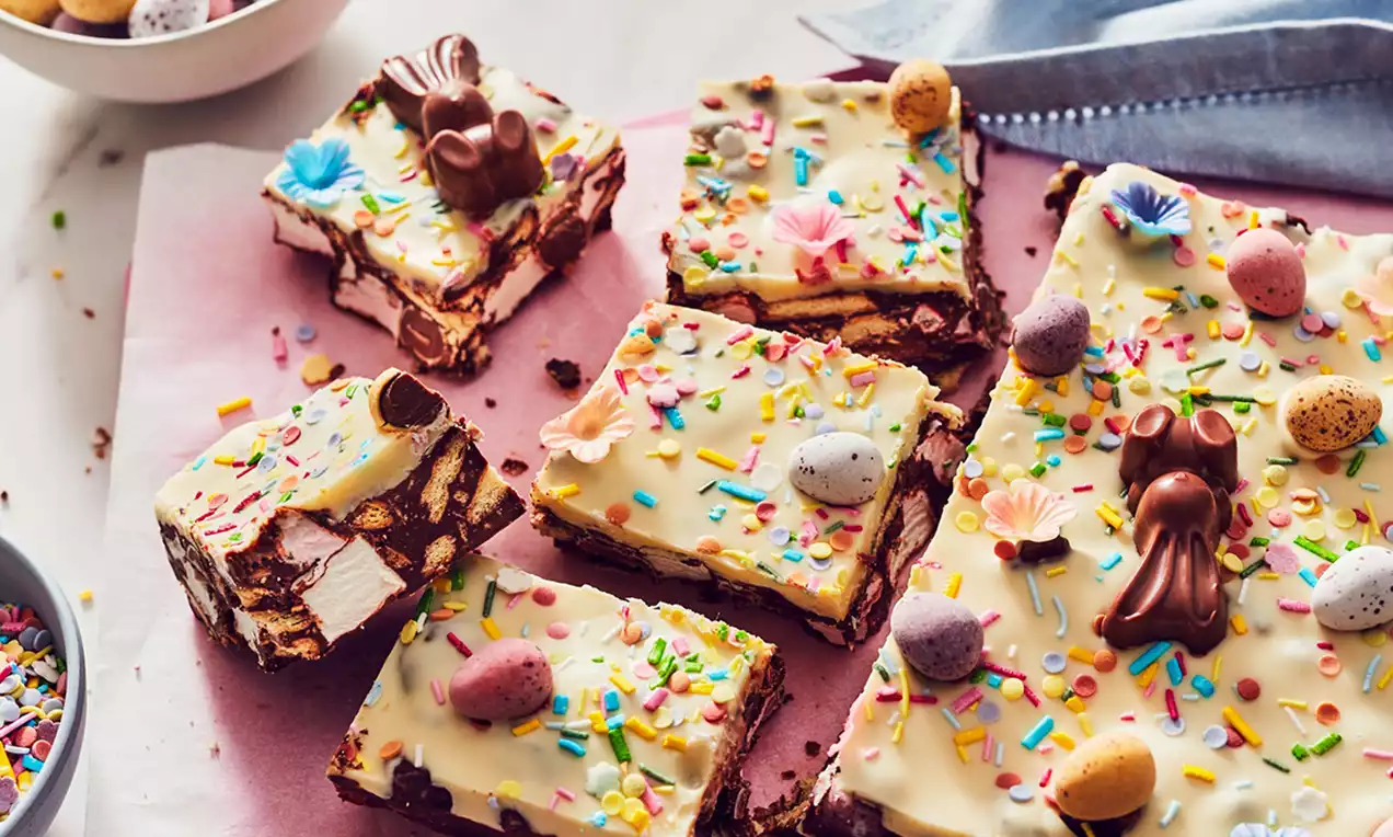 Bucăți pătrate de desert cu baza de ciocolată maro, strat de ciocolată albă, decor cu ouă colorate, iepurași și bombonele colorate pe hârtie roz