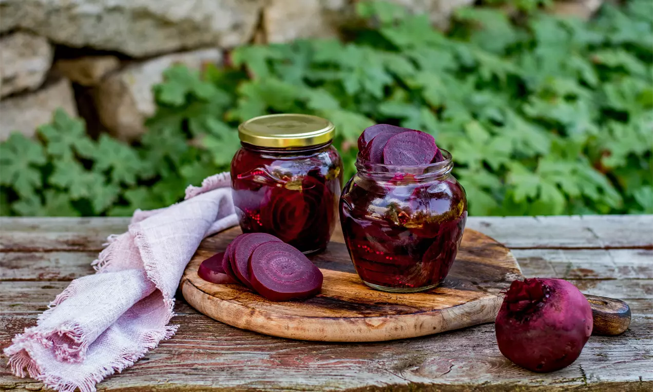 Тънки резени наситено червено цвекло в прозрачни буркани с орнаменти от марината, подредени върху дървена дъска на фон от зеленина
