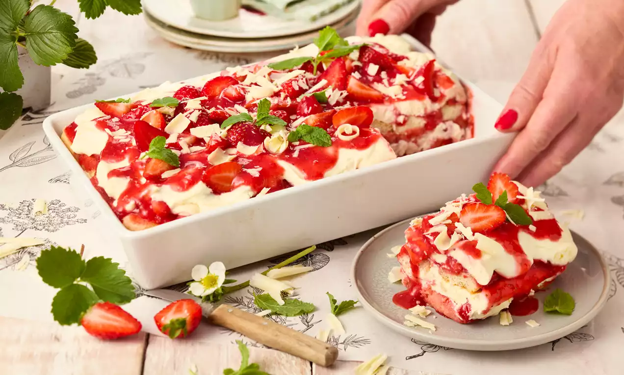 Schichtdessert in weißer Form mit roten Erdbeeren, heller Creme, Kräuterblättern und weißer Schokolade, serviert auf hellbraunem Tisch
