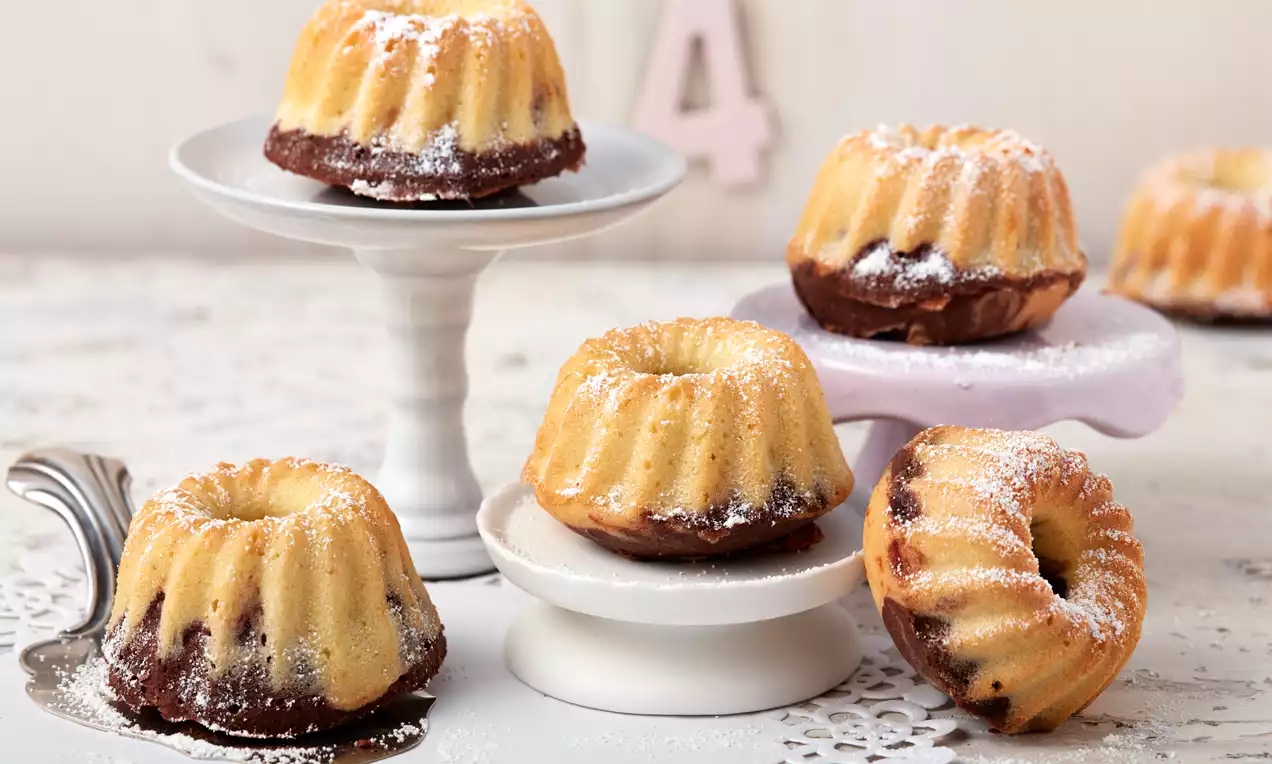 Mini-Marmor-Gugelhupfe mit goldgelber und schokoladener Marmorierung, bestäubt mit weißem Puderzucker auf weißen Kuchenständern und Spitzenunterlage