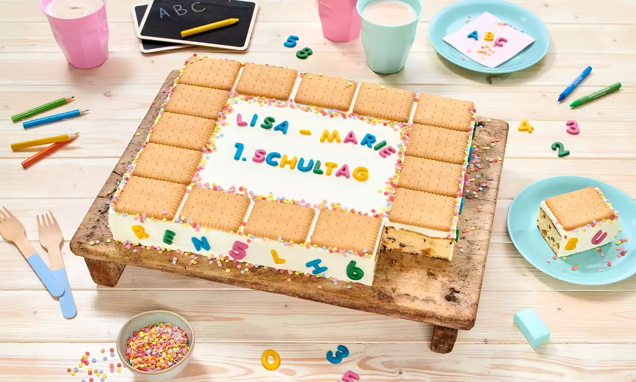 Pravokutna torta prekrivena bijelom kremom, ukrašena šarenim slovima, komadićima keksa na vrhu te posipom u boji na drvenom stalku