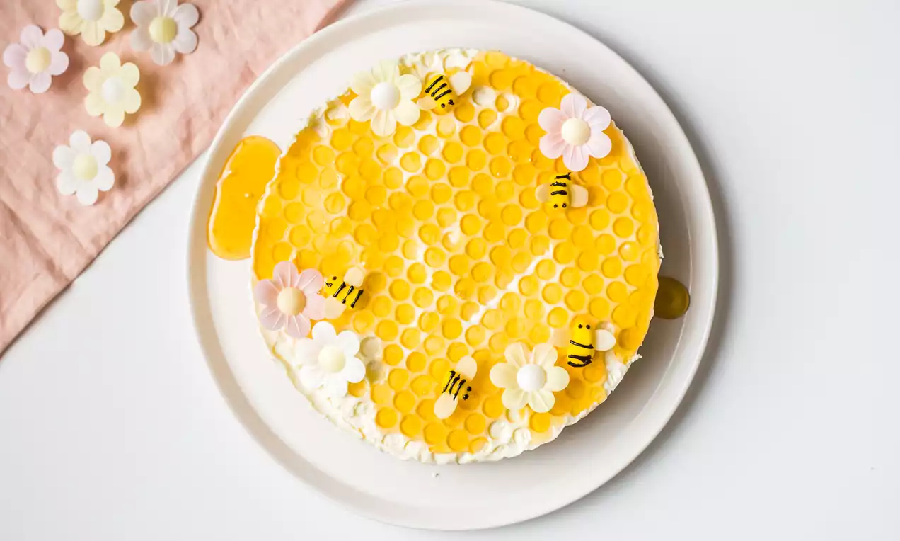Tarta blanca con capa amarilla brillante en patrón de panal, decorada con flores pastel, pequeñas abejas y miel sobre plato blanco