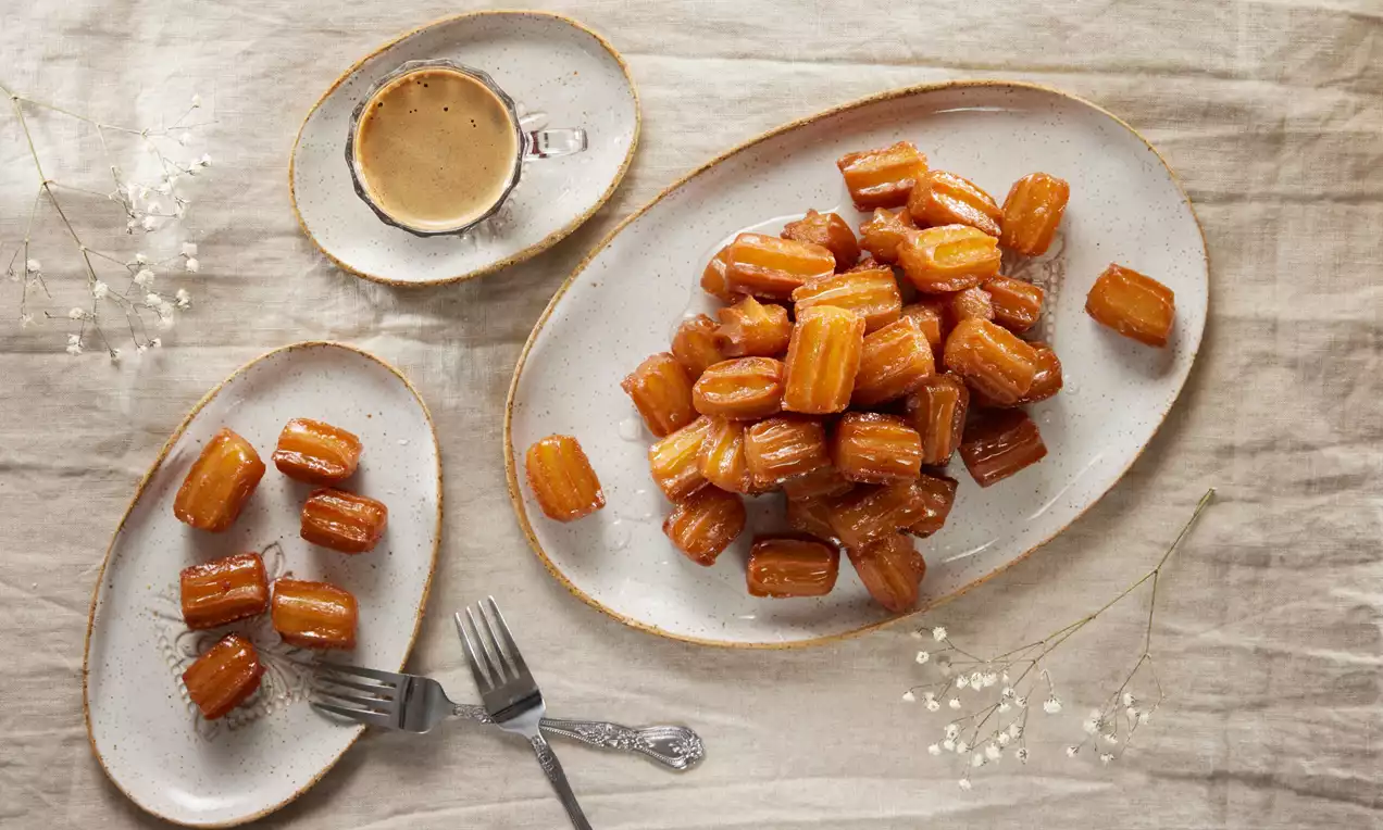 Altın renginde tulumba tatlıları parlak şerbetli, oval beyaz tabakta düzenlenmiş, yanında kahve fincanı ve çatal konumlandırılmış