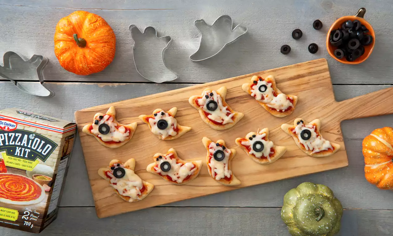 Ghost-shaped mini pizzas with melted cheese, red-orange tomato sauce, black olive eyes, arranged on a wooden board with pumpkins nearby