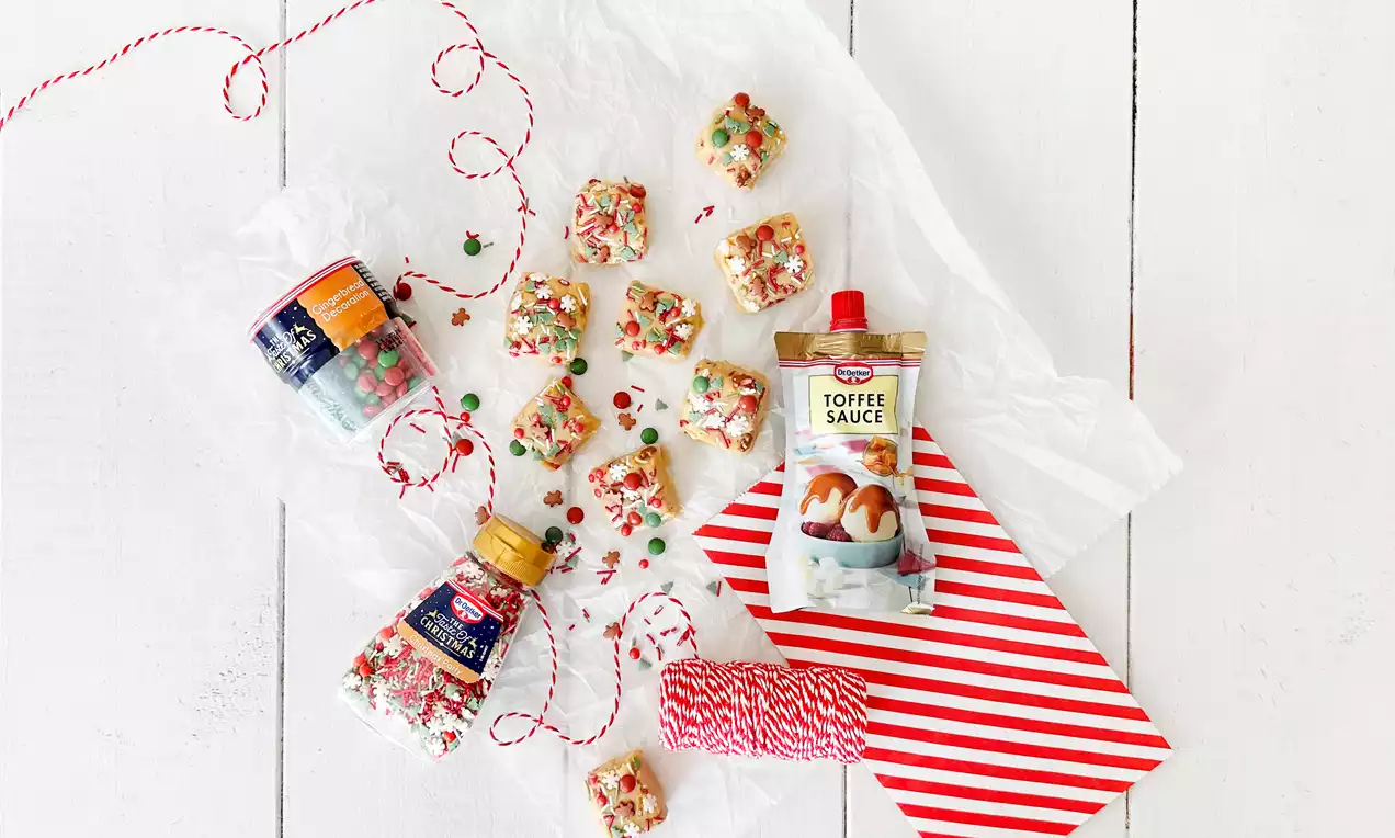 Neliönmuotoiset vaaleat fudge-palat koristeltu värikkäällä strösselillä ja aseteltu valkoiselle paperille punavalkoisen nauhan lomassa