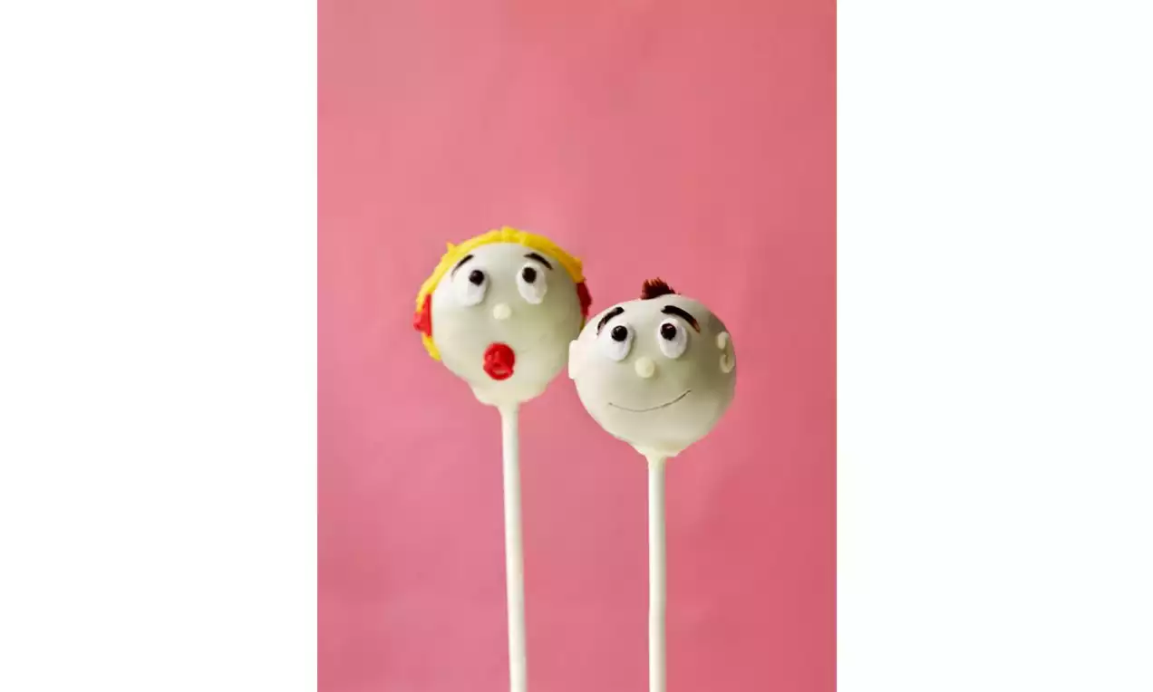 To hvite cake pops formet som ansikter med dekor i gul og brun glasur, rødt og svart pynt, på hvite pinner mot rosa bakgrunn