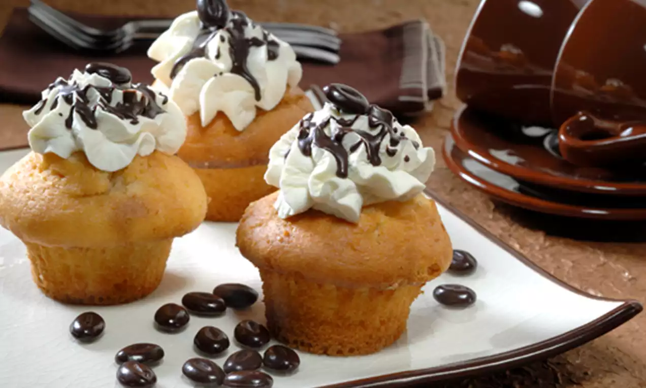 Drie lichtbruine muffins met slagroomtoef, chocolade drizzle en koffieboontjes op een wit bord, omgeven door losse koffiebonen en servies