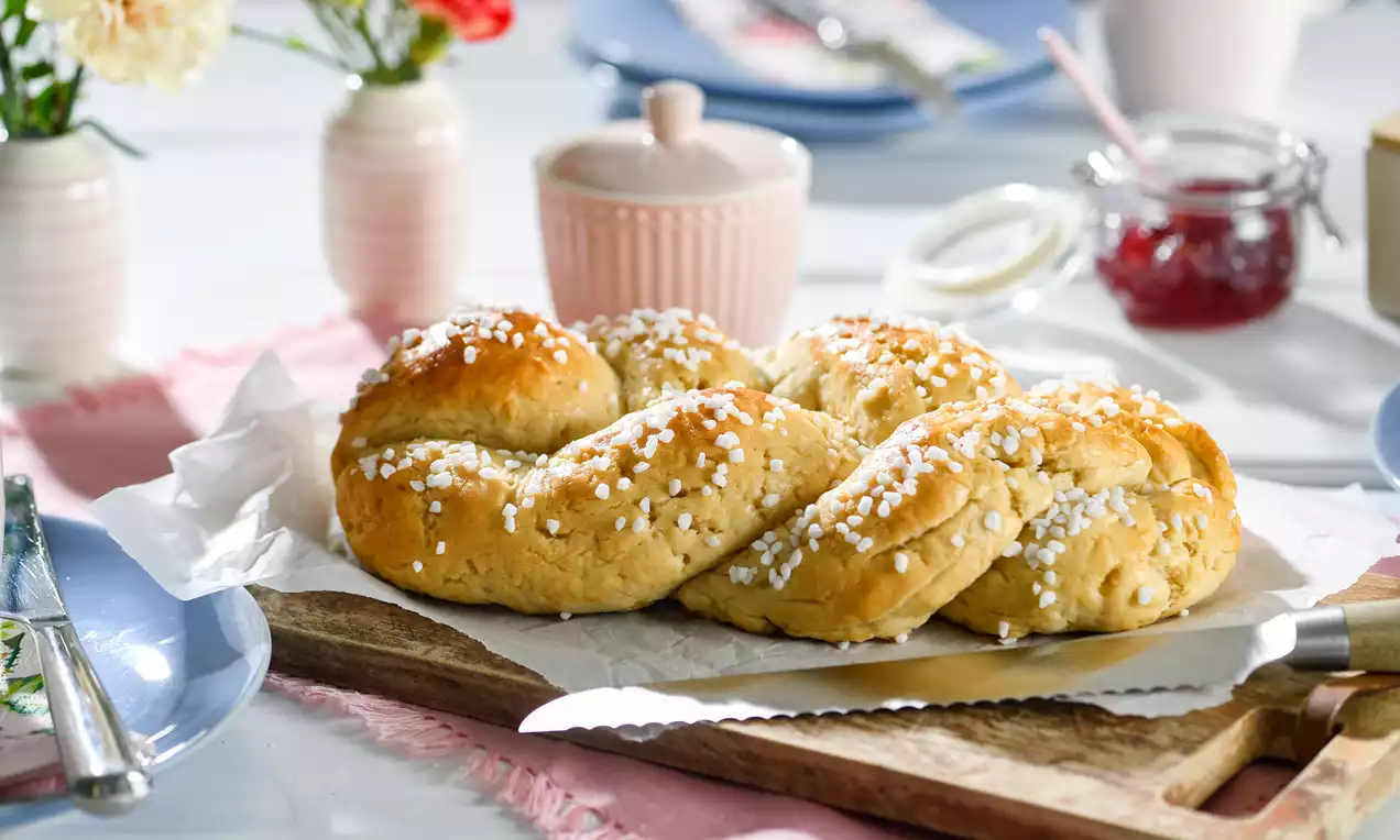 Goldbrauner glutenfreier Hefezopf mit grobem Hagelzucker bestreut auf weißem Papier auf einem Holzbrett, umgeben von Geschirr und Blumen