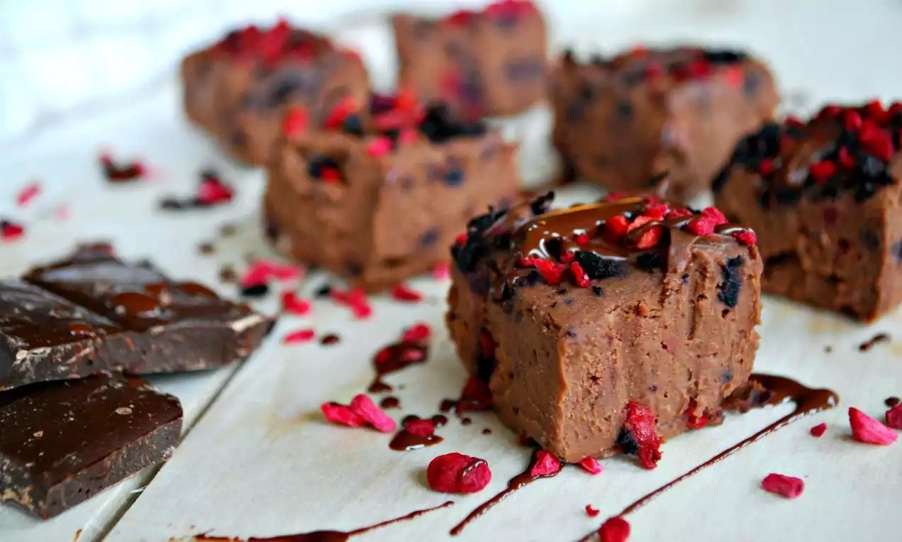 Browniebrune firkanter av sjokoladefudge med glatt overflate, toppet med røde bær og drypp av sjokoladesaus på hvitt pergamentpapir