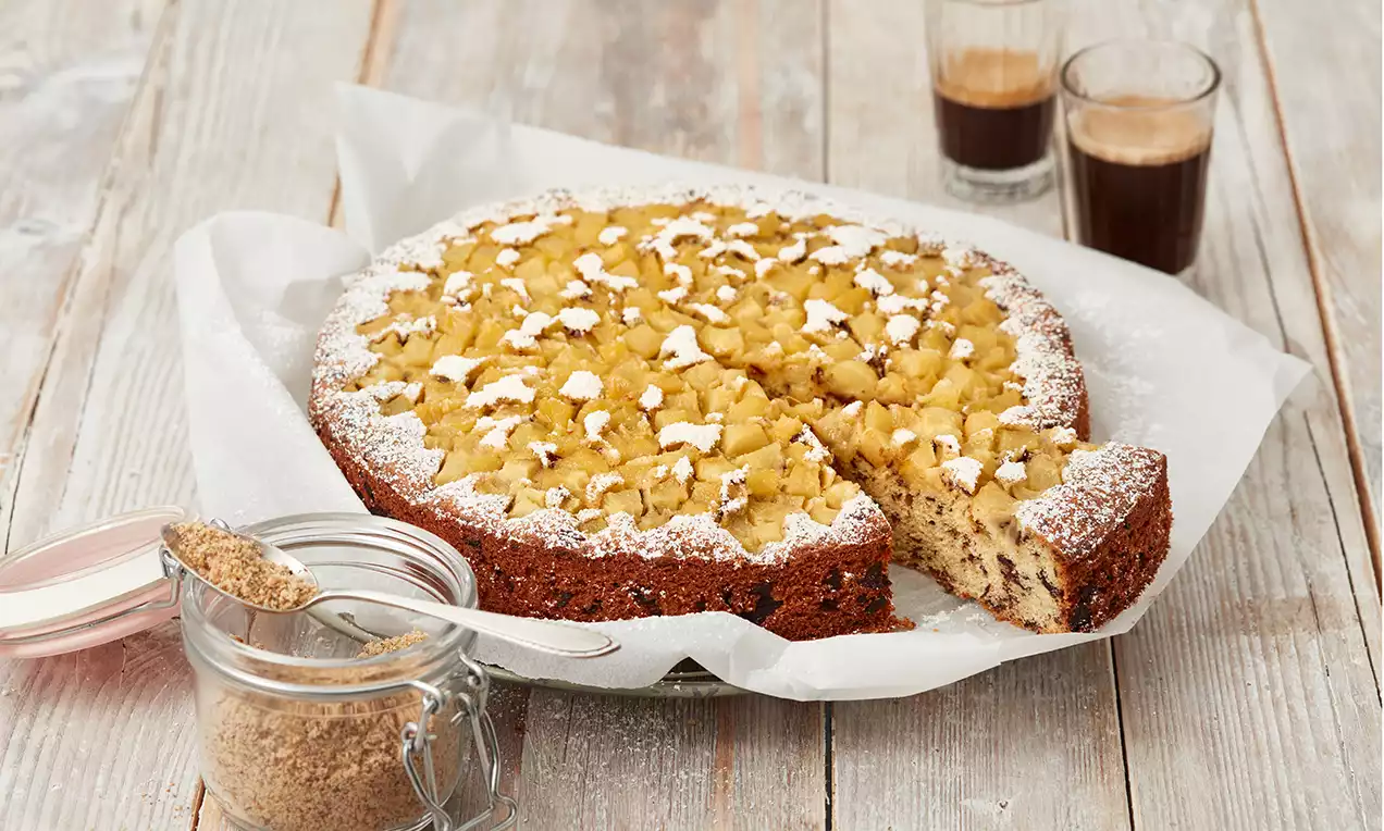 Saftiger Kuchen mit braunem Rand, goldgelber Birnenstückchen-Decke und Puderzucker auf Backpapier, daneben ein Glas mit braunem Zucker