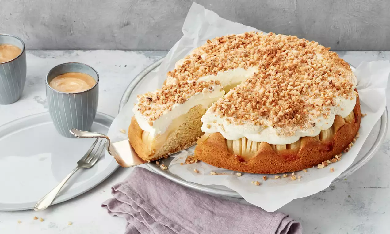 Herbstkuchen mit hellem Rührteig, frischen Apfelscheiben, luftiger Sahne und gerösteten Nusskrümeln auf weißem Teller mit lilafarbenem Tuch