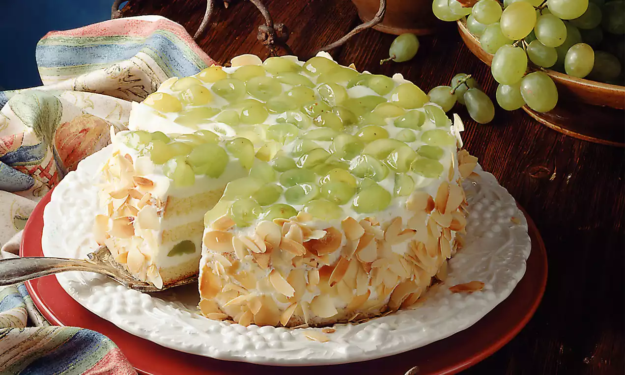 Kefirtorte mit glänzenden grünen Weintrauben auf hellem Guss, umrandet von goldbraunen Mandelblättchen auf einem weißen Teller