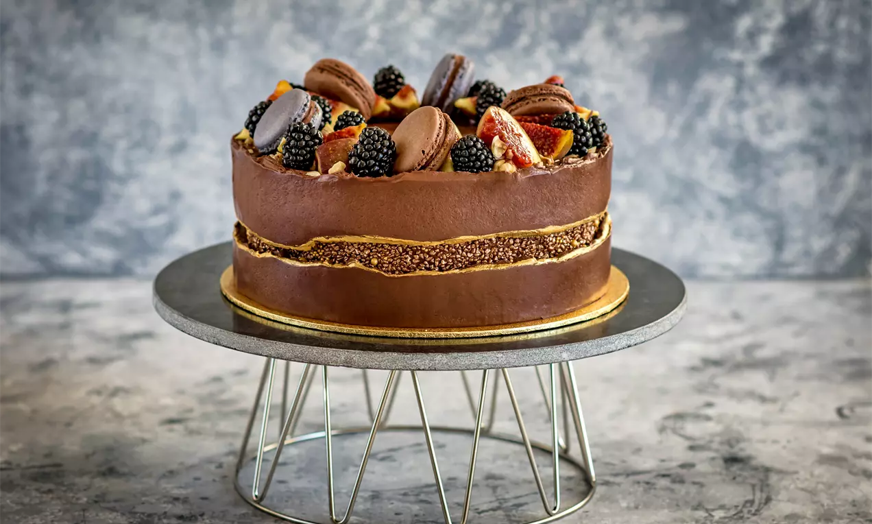 Шоколадова торта с лъскава глазура, поръсена с горски плодове, макарони и резенчета портокал, върху тъмна поставка